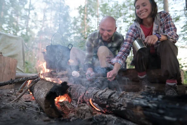 Amici felici friggono marshmallow su un fuoco, sopra la fiamma di cui pende un bollitore, una donna con una tazza in attesa di una bevanda calda. — Foto Stock