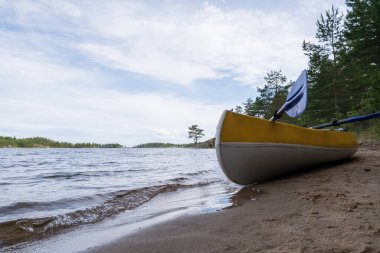 Kürekli kano, bir gölün kıyısındaki kumlu bir sahilde çam ağaçlarının ve gökyüzünün arka planına park edilmiştir. Etkin dinlenme.