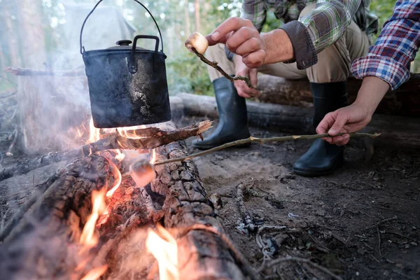 Due amici friggono dolci marshmallow su un fuoco con un bollitore sulla fiamma, in una giornata estiva, in un campeggio, nella foresta. — Foto Stock