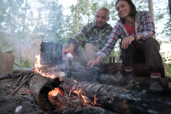 Soddisfatti, deliziosi amici friggono un delizioso marshmallow su un fuoco, sulla cui fiamma pende un bollitore con una bevanda calda.. — Foto Stock