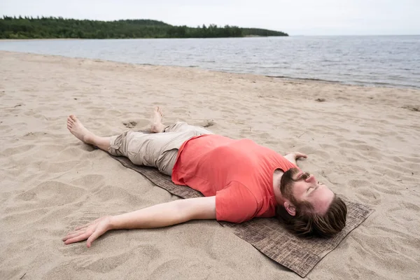 Skäggig man gör yoga, han gör Savasana övningen, slappnar av i en likställning liggande på en matta, på en sandstrand vid sjön. — Stockfoto