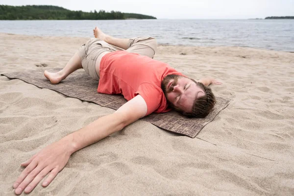 Uomo barbuto pratica yoga, esegue un esercizio sdraiato su un tappeto, su una spiaggia di sabbia vicino al lago, in una giornata estiva. — Foto Stock
