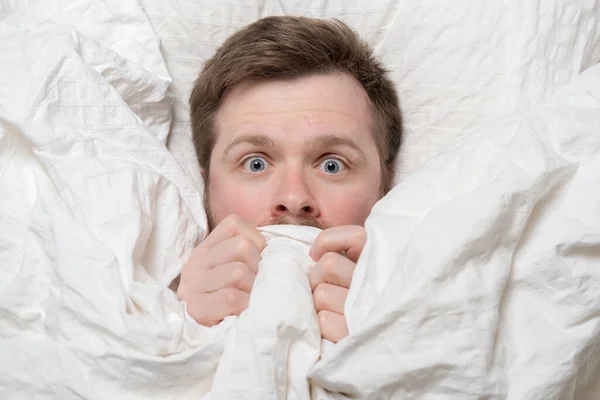 Nightmare. Caucasian man is scared, he hides under the covers and looks with big eyes, lying in bed. Psychological health concept. — Stock Photo, Image