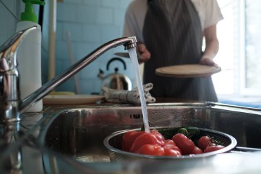 Lavabodaki kevgire akan suyun altında taze sebzeler, bıçaklı ve kesme tahtası olan bir eşin arka planına karşı..
