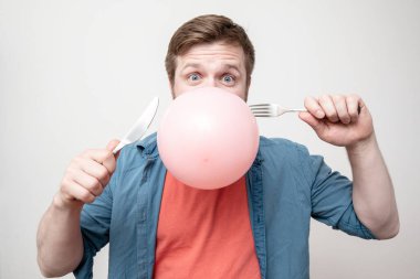 Ağzında bir balon tutan komik bir adam çatal ve bıçakla onu patlatacak. Beyaz arkaplan.