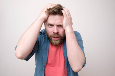 Adam stresli ya da şiddetli bir baş ağrısı çekiyor, elleriyle başını tutuyor ve şaşkınlık içinde kameraya bakıyor. Beyaz arkaplan.