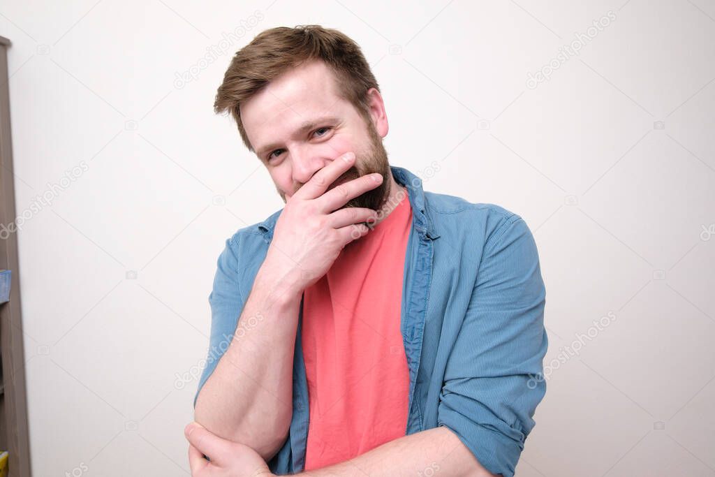 Shy, happy man smiles while covering his mouth with his hand. White background.