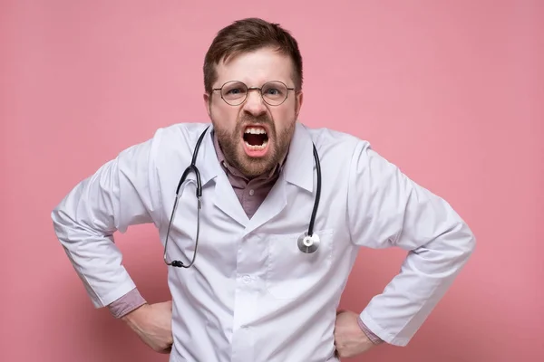 Disgruntled male doctor wearing glasses, a white coat and a stethoscope around neck screams angrily while looking into the camera. — Stock Photo, Image