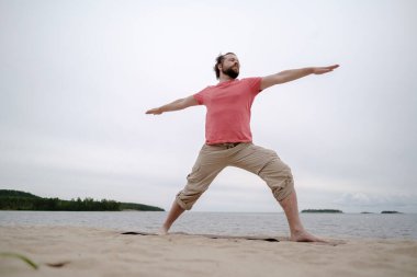 Kafkasyalı adam yoga yapıyor, virabhadrasana egzersizi yapıyor, halının üzerinde duruyor, göl kıyısındaki kumlu bir sahilde..