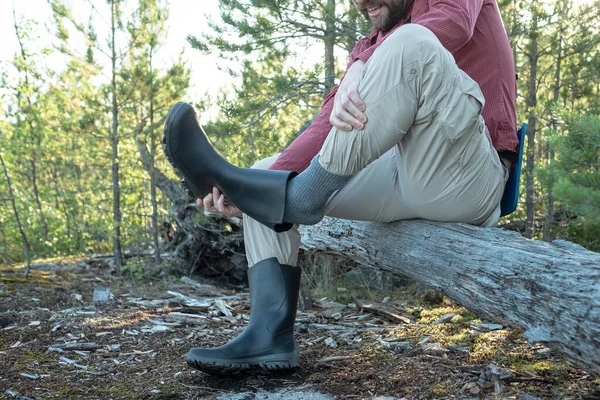 Man in hiking clothes sat on an old log in the woods, declines rubber boot that shakes dust from it. Active lifestyle concept.