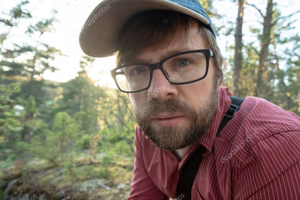 Bearded man in glasses and a cap resting in the forest, after a long walk. He looks intently and thoughtfully. Close-up.