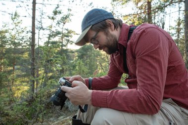 Fotoğrafçı dinleniyor, elinde bir kamera tutuyor ve ormanda yürürken çekilen fotoğraflara ilgiyle bakıyor..