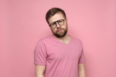 Şüpheli, sakin, gözlüklü, düşünceli, gözlerini kısarak kameraya bakan beyaz bir adam. Pembe arkaplan.
