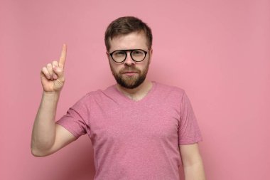 Sakallı, gözlüklü, işaret parmağı takan sakin bir adam kameraya bakar. Pembe arkaplan.