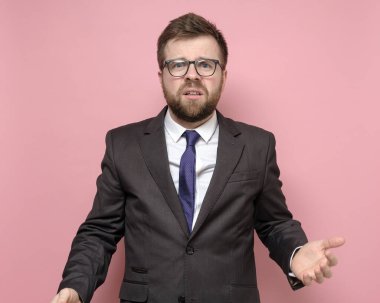 Takım elbiseli bir adam şaşkına döner, elleriyle bir jest yapar ve öfkeyle kameraya bakar. Pembe arkaplan.