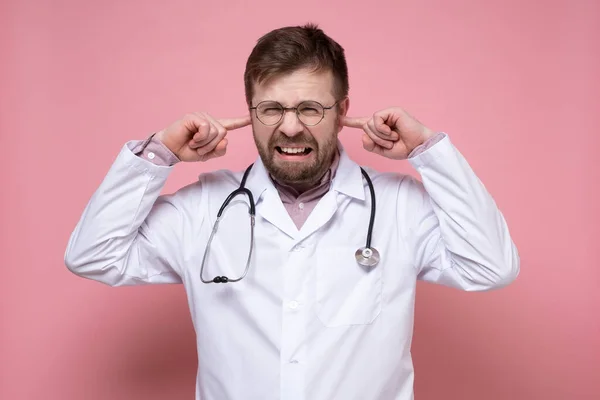 Doctor with a stethoscope around neck is stressed, he is annoyed by the noise, he covers ears with fingers and bares teeth.