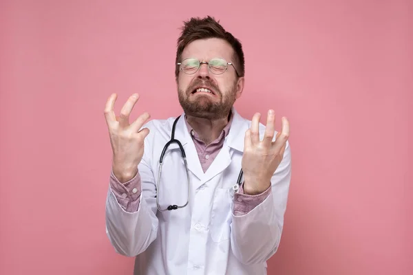 Steteskop boynuna dolanmış bir doktor dişlerini öfkeyle gösteriyor ve elleriyle bir jest yapıyor. Pembe arkaplan. — Stok fotoğraf