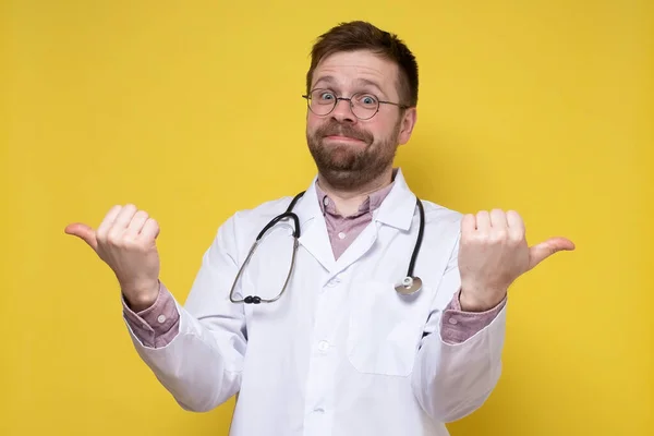 Médico engraçado com um estetoscópio em torno do pescoço aponta polegares em duas direções e olha para a câmera de surpresa, sorrindo. — Fotografia de Stock