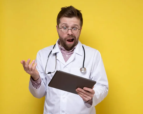 Boynunda steteskop olan genç bir doktor elinde bir tablet tutuyor ve şaşkınlıkla ona bakıyor. Sarı arkaplan. — Stok fotoğraf