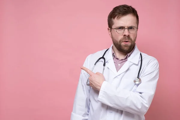 Serious young doctor with a stethoscope around neck points finger to the side and looks anxiously at the camera. Copy space. — Stock Photo, Image