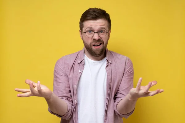 Perplexed Caucasian bearded man in glasses makes a gesture with hands and looks inquiringly into the camera. Yellow background. — Stock Photo, Image