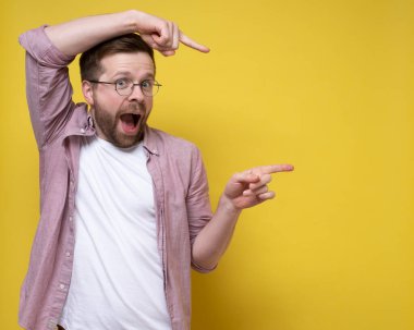 Komik adam işaret parmağını bir şeye doğrultuyor, sıra dışı bir pozisyonda duruyor ve mutlu bir şekilde kameraya bakıyor. Boşluğu kopyala. 