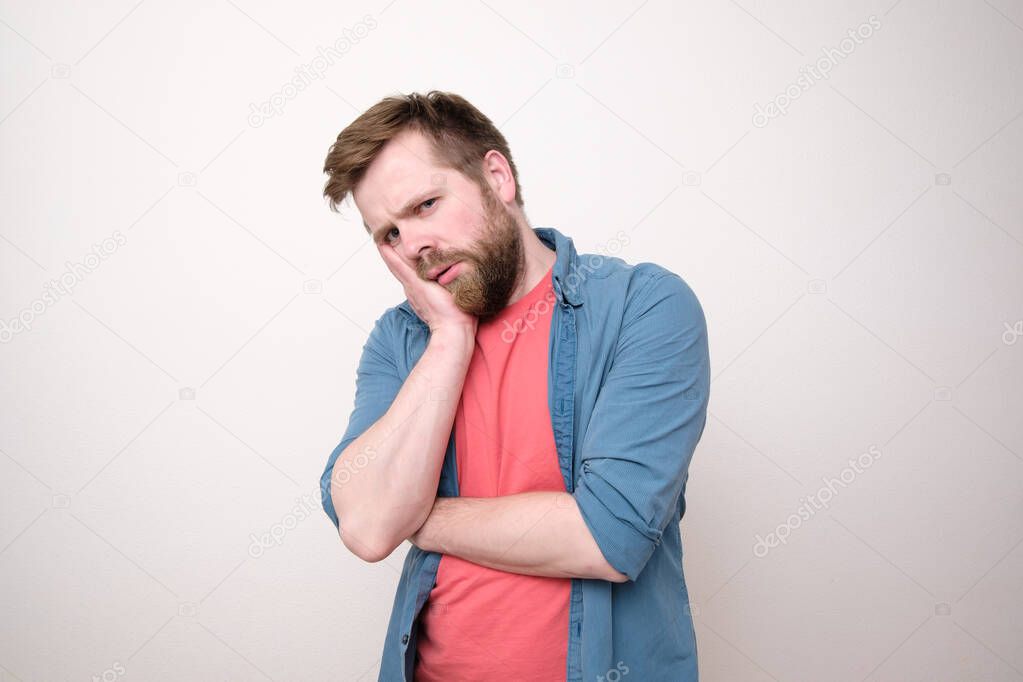 Caucasian man is upset or has a toothache, he holds hand on cheek and looks sad. White background.