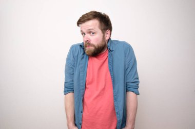 Şaşkın sakallı bir adam düşünceli ve şaşırmış görünüyor, ellerini ceplerine sokuyor ve omuzlarını kaldırıyor. Beyaz arkaplan.