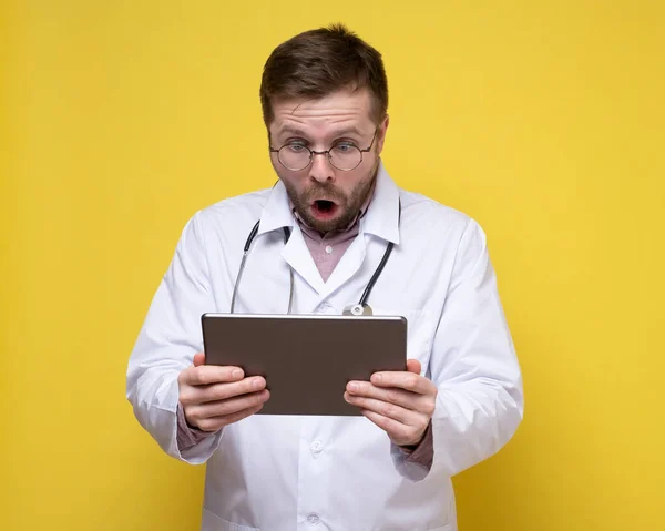 Korkmuş, şok olmuş bir doktor, ağzı açık bir tablete ve kocaman gözlere bakar. Sarı arkaplan. — Stok fotoğraf
