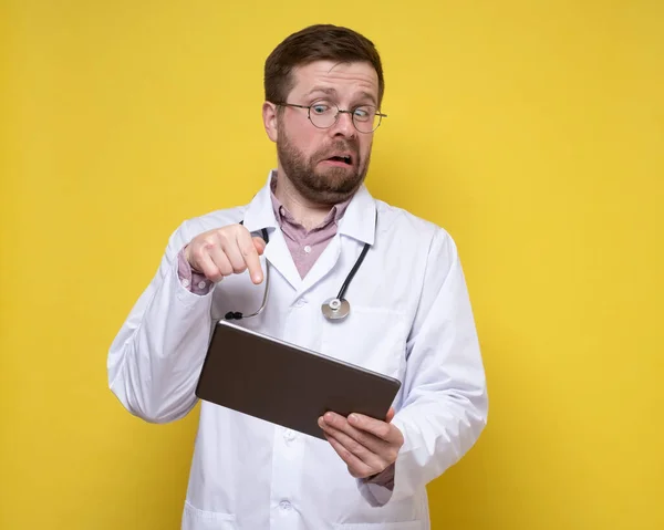 Şaşırmış doktor elinde bir tablet tutuyor ve heyecanlı bir şekilde ekranı işaret ediyor. Sarı arkaplan. — Stok fotoğraf