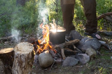 Kamp ateşi, bir çaydanlığın asılı olduğu alevin üzerinde ve yemek hazırlanıyor, arka planda bir erkek bacağı, kampta. 