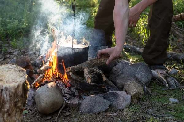 L'homme met du bois de chauffage sur un feu de camp, sur lequel il cuisine, dans le camp. Tourisme et mode de vie actif. — Photo