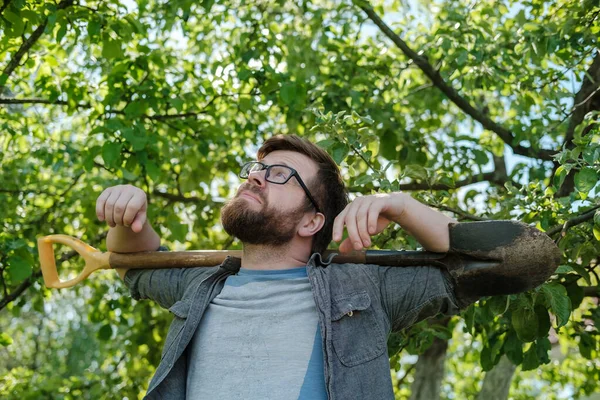 Bearded man in glasses holds a shovel on shoulders and looks up, dreamily, thoughtfully, against the backdrop of a garden. — Stock Photo, Image