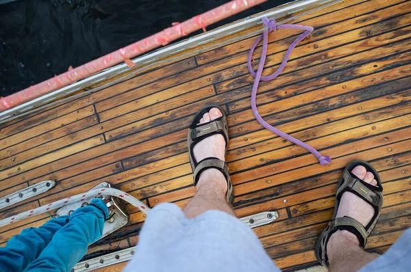 Teknenin ahşap güvertesindeki sandaletli erkek ayakları karanlık suyun arka planında duruyor. Üst görünüm. — Stok fotoğraf