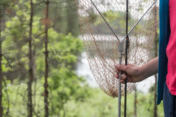 Hand houdt een net voor het trekken van vis uit het water, tegen de achtergrond van een meer en bomen, op een zomerdag. — Stockfoto