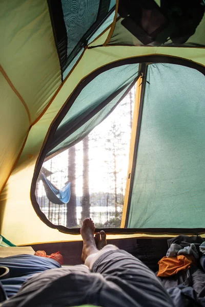 O homem está relaxado em uma tenda, em um acampamento, contra o pano de fundo de uma rede suspensa, um lago e uma floresta. Estilo de vida. — Fotografia de Stock