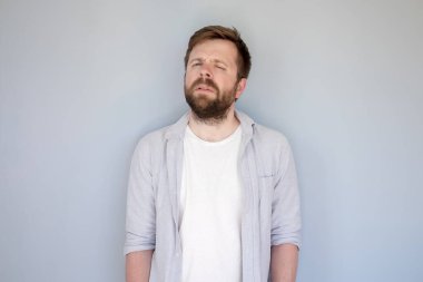 Gri arka planda dikilirken, günlük giysiler içinde beyaz, sakallı adam şüpheli görünüyor..