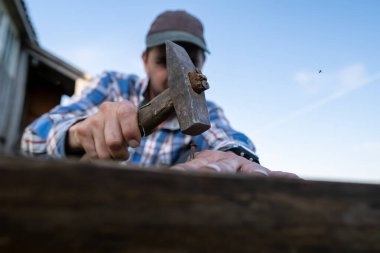 Marangoz elleri, mavi gökyüzüne karşı bir köy avlusundaki tahtaya çivi çakarak bir bank yapar..
