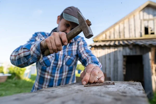 Odaklanmış marangoz, bir yaz günü arka planda bir ahır olan bir köy avlusunda bir tahtaya çivi çakıyor..