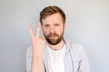  Gömlekli ciddi sakallı bir adam parmaklarıyla zaferin sembolünü gösterir. Gri arkaplan.