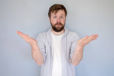 Şaşkın sakallı, gömlekli bir adam, şok oldu, ellerini yana doğru kaldırdı ve şaşkın şaşkın baktı. Gri arkaplan.