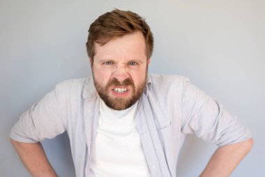 Beyaz sakallı adam öfkeli ve hoşnutsuz, dişlerini gösteriyor ve kızgın görünüyor. Gri arkaplan.