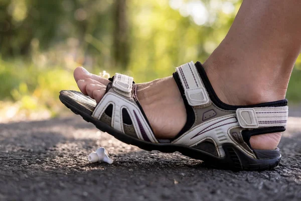 Frau Fuß in Sandalen Schritte auf einem schäbigen drahtlosen Kopfhörer, der auf einem asphaltierten Bürgersteig liegt, im Freien, vor einem verschwommenen Hintergrund. — Stockfoto