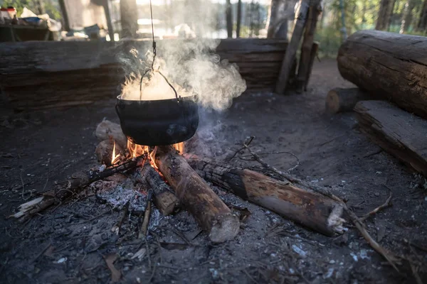 Bowler com vapor escapando paira sobre uma fogueira, em que a comida é preparada, ao pôr do sol, contra o pano de fundo de um lago. — Fotografia de Stock