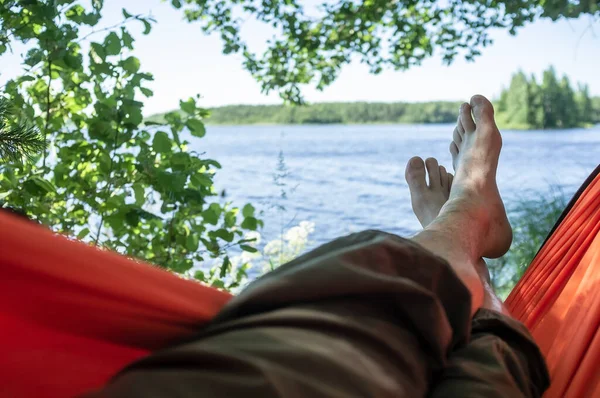 Carefree barefoot man relaxes lying in a hammock in the shade of trees, with a magnificent view of the lake, on a summer day. — Stock Photo, Image
