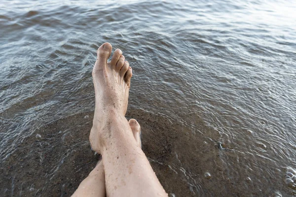 Мокрі жіночі ноги у воді, на піщаному пляжі. Жінка відпочиває, сидячи біля озера, в літній день. Стиль життя . — стокове фото