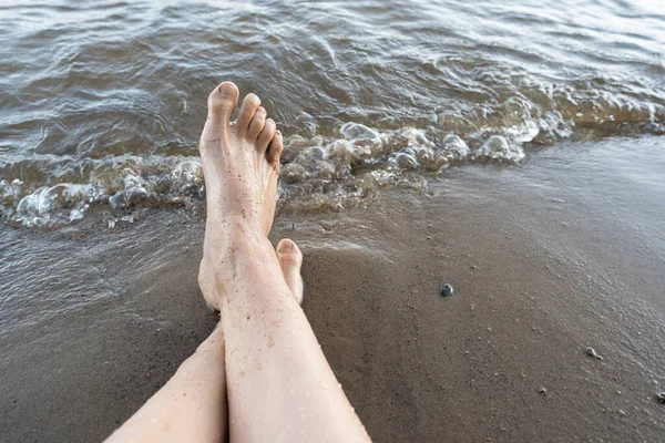 Pies femeninos mojados en una playa de arena cerca del agua. La mujer descansa sentada junto al lago, en un día de verano. Estilo de vida. — Foto de Stock