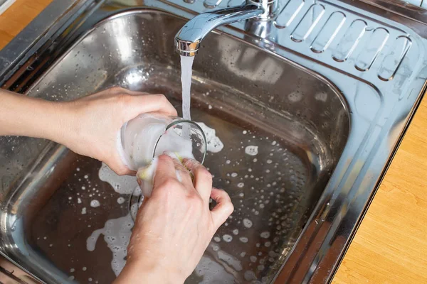 Frauenhände waschen ein Glas unter fließendem Wasser in der Küche mit Spülmittel und Schwamm. Hausarbeit. — Stockfoto