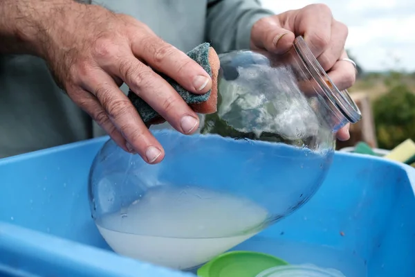 Männliche Hände waschen ein schmutziges Glasgefäß mit einem Schwamm, in einem Plastikbecken, in einem Dorfhof, im Freien. Ländliche Traditionen. — Stockfoto