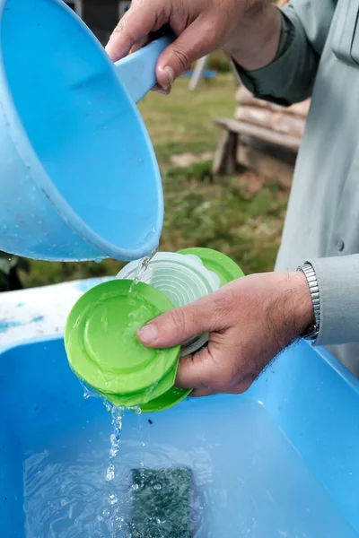 Männliche Hände gießen sauberes Wasser aus einer Schöpfkelle auf Plastikdeckel, um das Waschmittel auf einem Bauernhof im Freien abzuspülen. Ländliche Traditionen. — Stockfoto